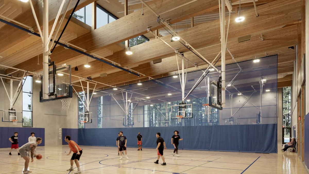 A gym with exposed wood and wooden ceilings is full of light as people play basketball.