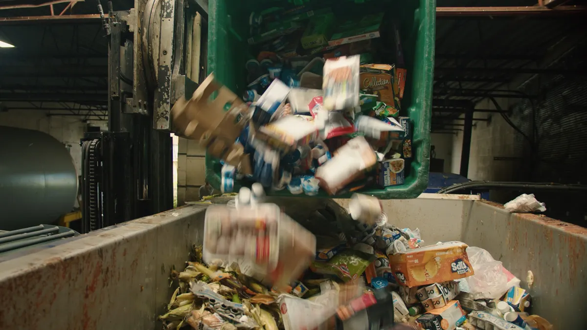 A large bin full of packaged, uneaten food is tipped into a dumpster by a machine inside a facility.