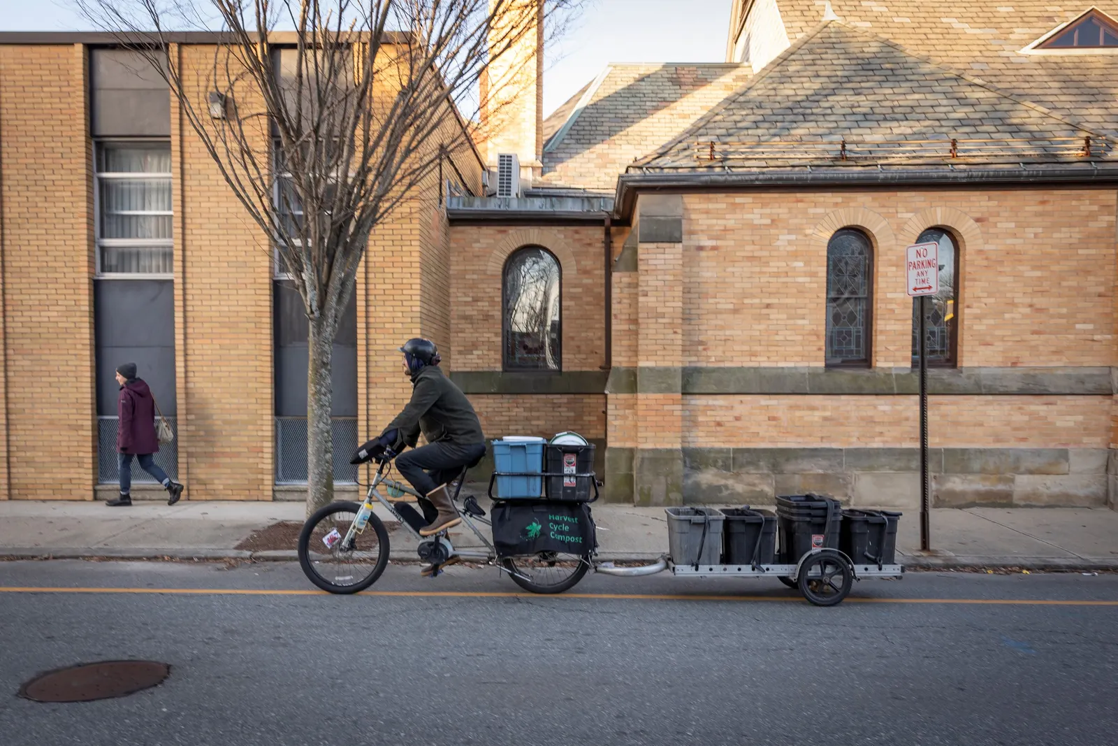 A man wearing a helmet pedals a bicycle down a city street. The bicycle is carrying several plastic buckets on a trailer, and has saddle bags that read "Harvest Cycle Compost."