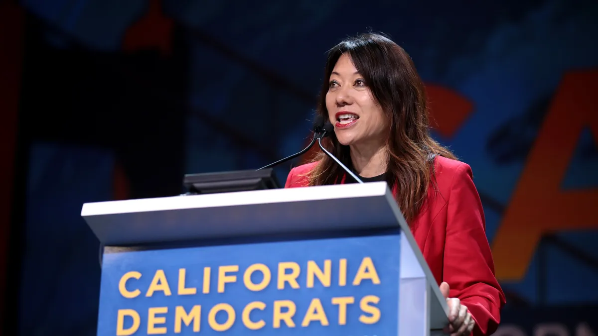 California Treasurer Fiona Ma stands in a red jacket behind a lectern with the words "California Democrats State Convention" on the front.