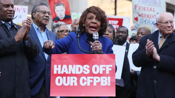 Rep. Maxine Waters speaks into a microphone with a crowd of people behind her, some holding signs. A sign in front of her podium reads "Hands off our CFPB!"
