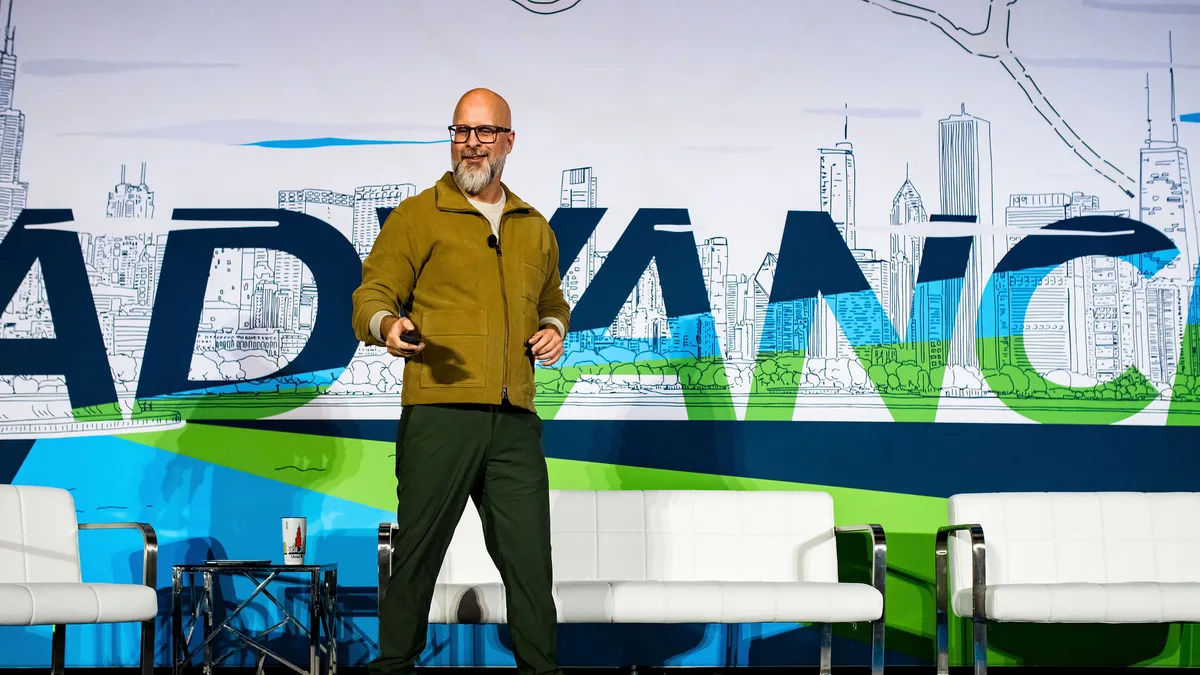 A person in glasses and a long sleeve shirt stands on a stage in front of the word "Advance"