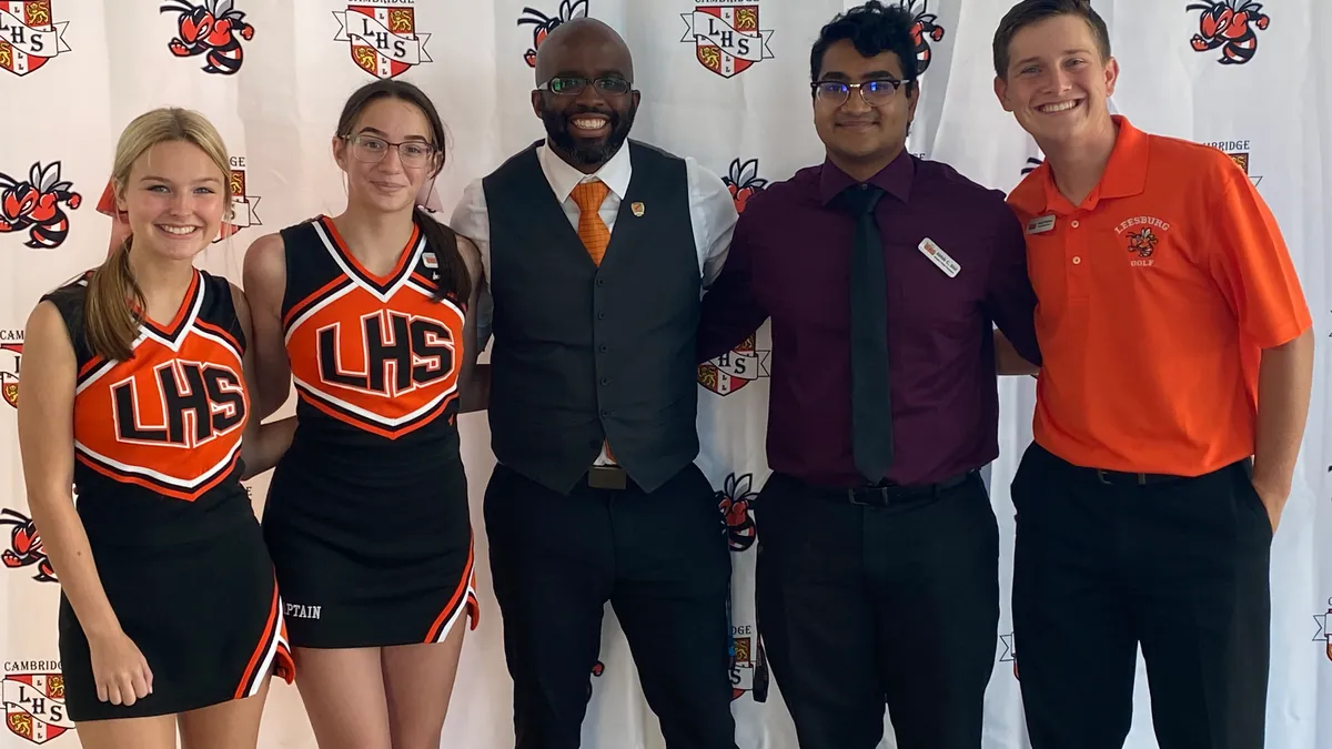 An adult and four high school students stand in a row in front of a white backdrop that had logos on it.