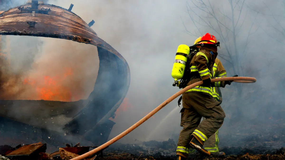 Two people in firefighting suits carry a hose outside. Fire and smoke is billowing in front of them.