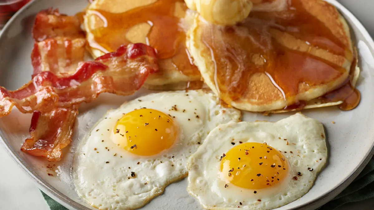 A photo of a plate with two fried eggs, two slices of bacon and two pancakes with syrup.