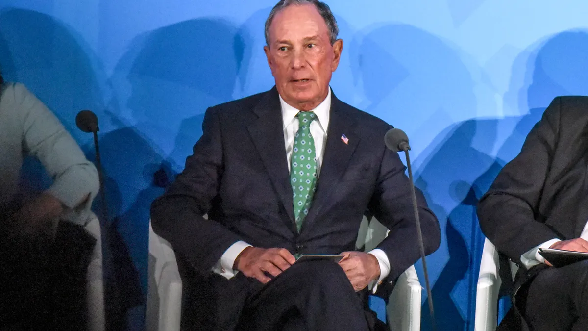 Michael Bloomberg is seated in the center of the frame wearing a black suit with an American flag pin on the left lapel, photo right, wearing a white shirt and green tie.