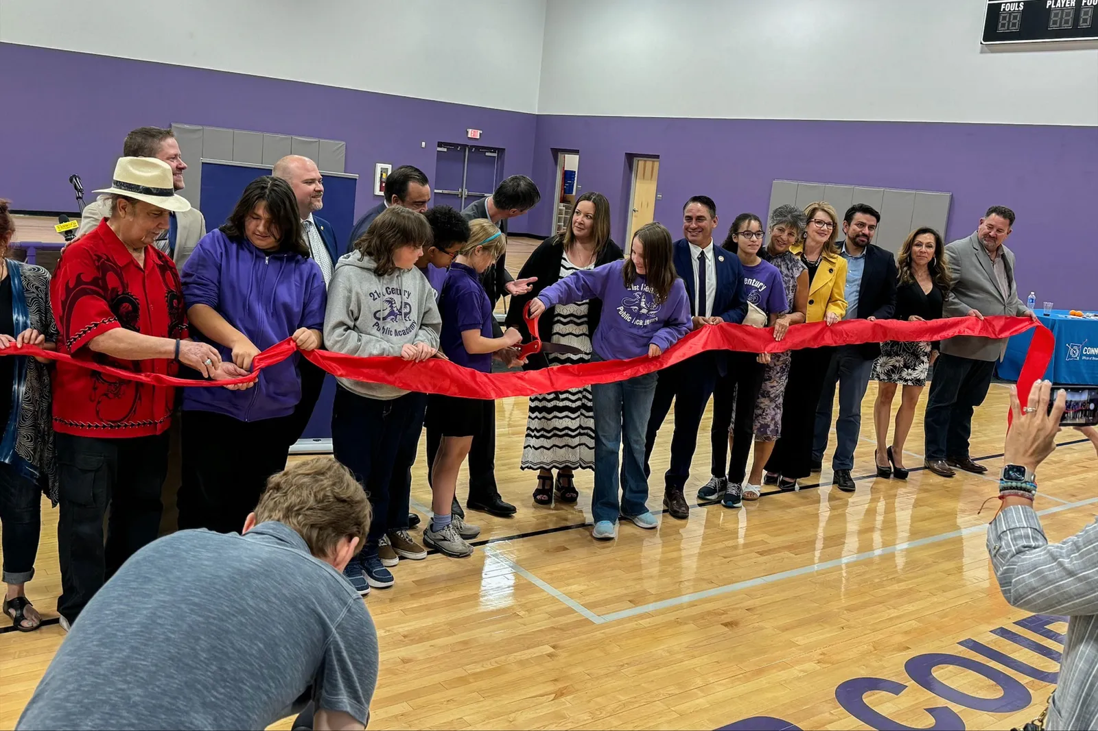 People are lined up in a row behind a long red ribbon that they are holding. They are in a school gym that has purple walls. A person with a camera is facing the row of people.