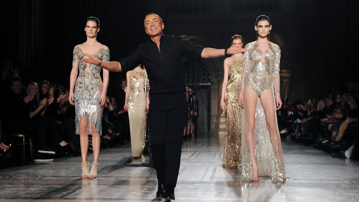 Julien Macdonald walks the runway with models after his show. The designer wears all black and stretches his arms out in gratitude to the audience, while models stand behind him in metallic outfits.