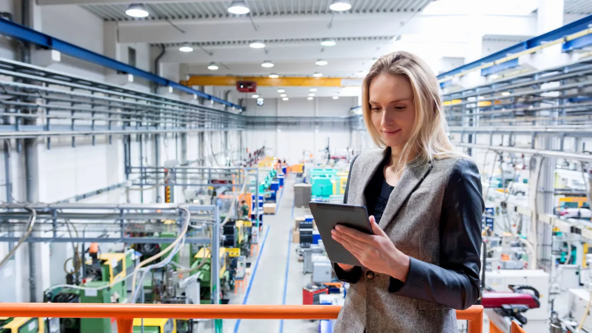 Horizontal color image of blond business female worker standing on balcony on top of large factory, holding digital tablet and examining the production online. Focus on attractive businesswoman, futur