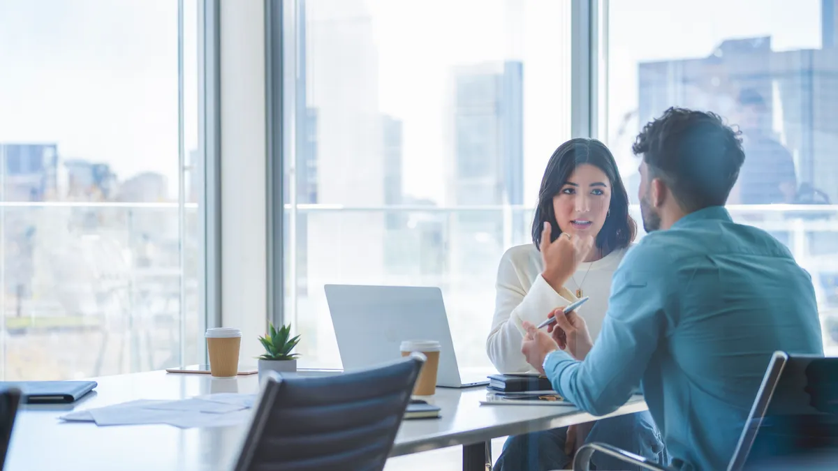 Two workers taking in a conference room. There is a laptop on the table in the room.