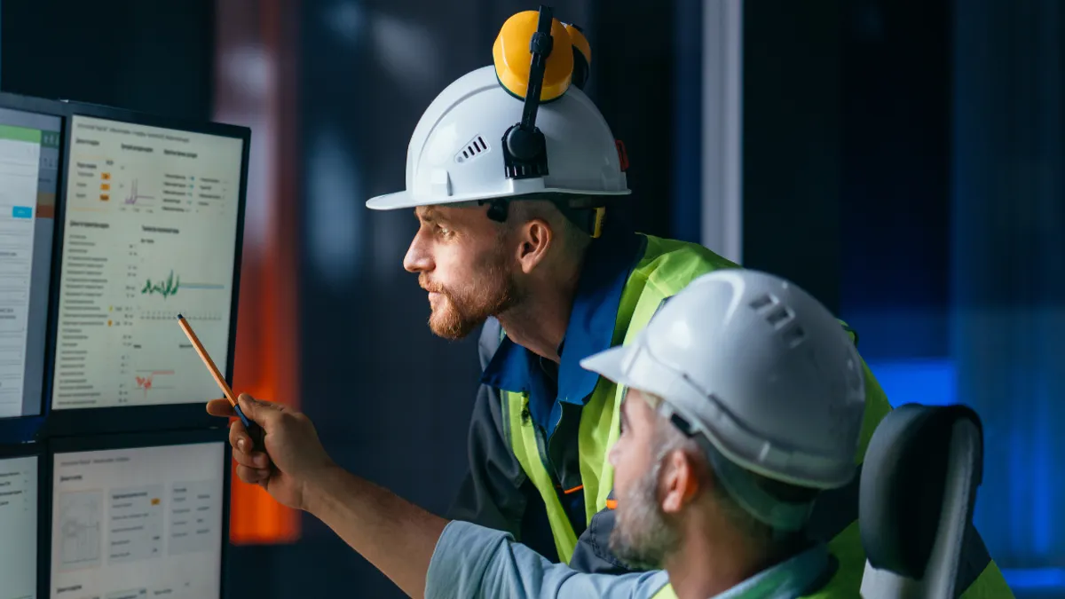 Two engineers in safety helmets and high-visibility vests reviewing data and graphs on large computer monitors inside a control room.