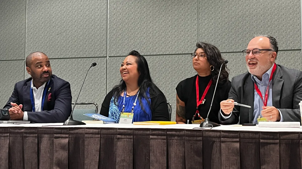 Four people sit at a wide desk on stage, speaking to an audience that is out of frame.