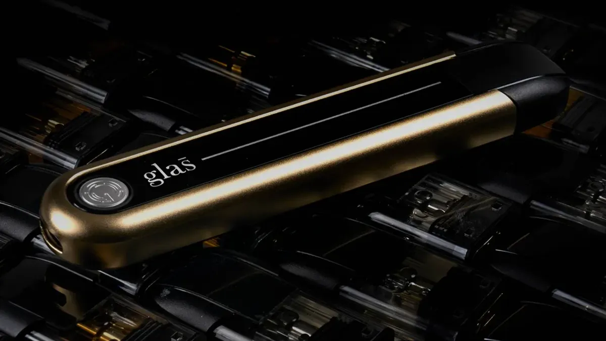 A vaping device lies on a bed of vaping liquid pods. The device is gold and black with a small silver seal and says "Glas" on one end.