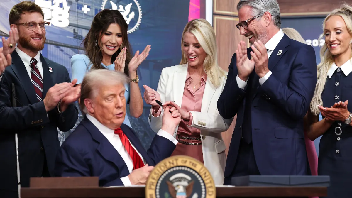 Man with a pen in hand at a desk with President of the United States seal with people standing around him clapping