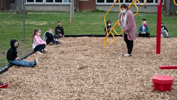 Young students sit on the ground outside near playground equipment. A teacher is standing looking at the children. Everyone is wearing face masks.