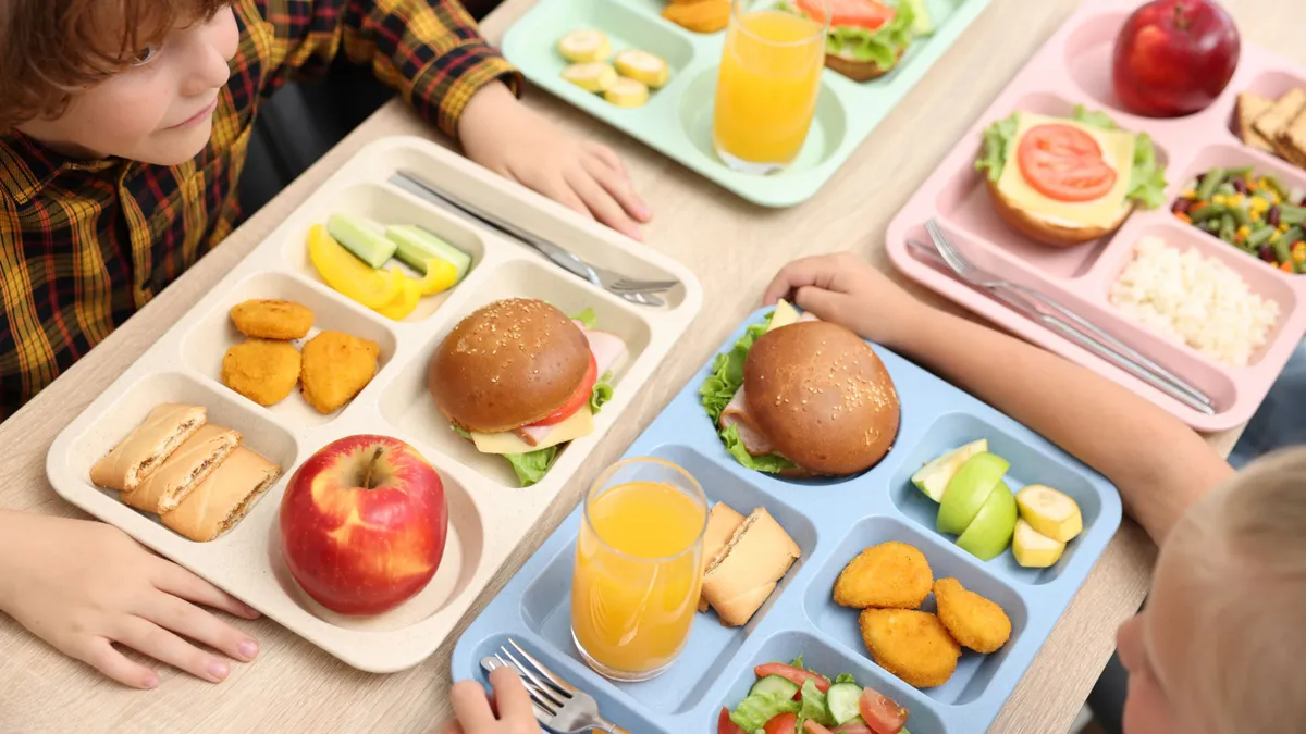 Students sit at a long table with four colorful lunch trays sitting close together.