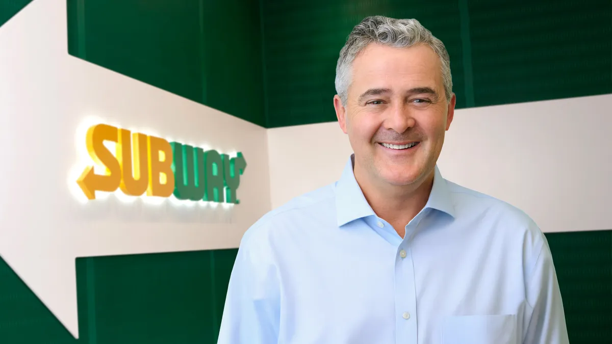 A smiling person stands in front of a green background with a yellow "Subway" logo visible.