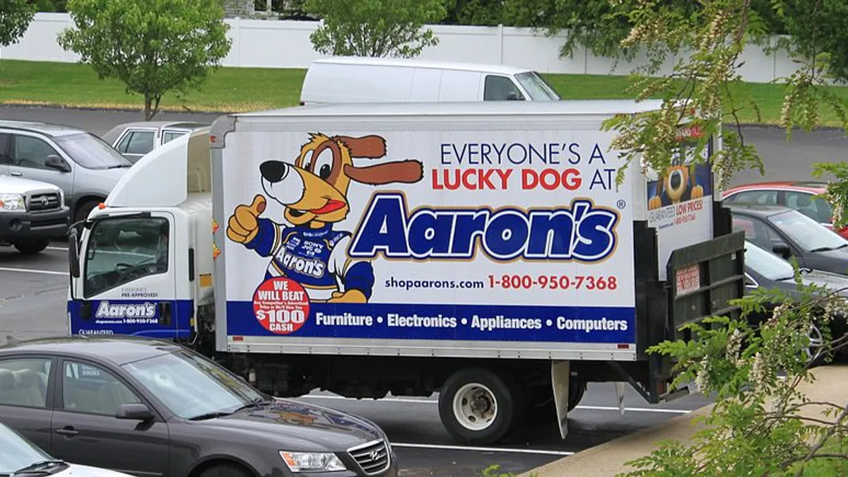 A delivery truck with the Aaron's logo and a cartoon dog is shown in a parking lot amogst other vehicles.