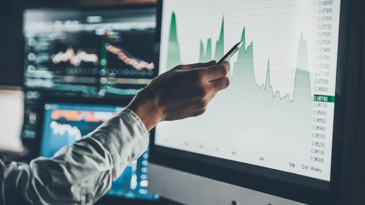 A person points a pen at a computer with a green line graph. A blurred computer in the background shows other graphs.