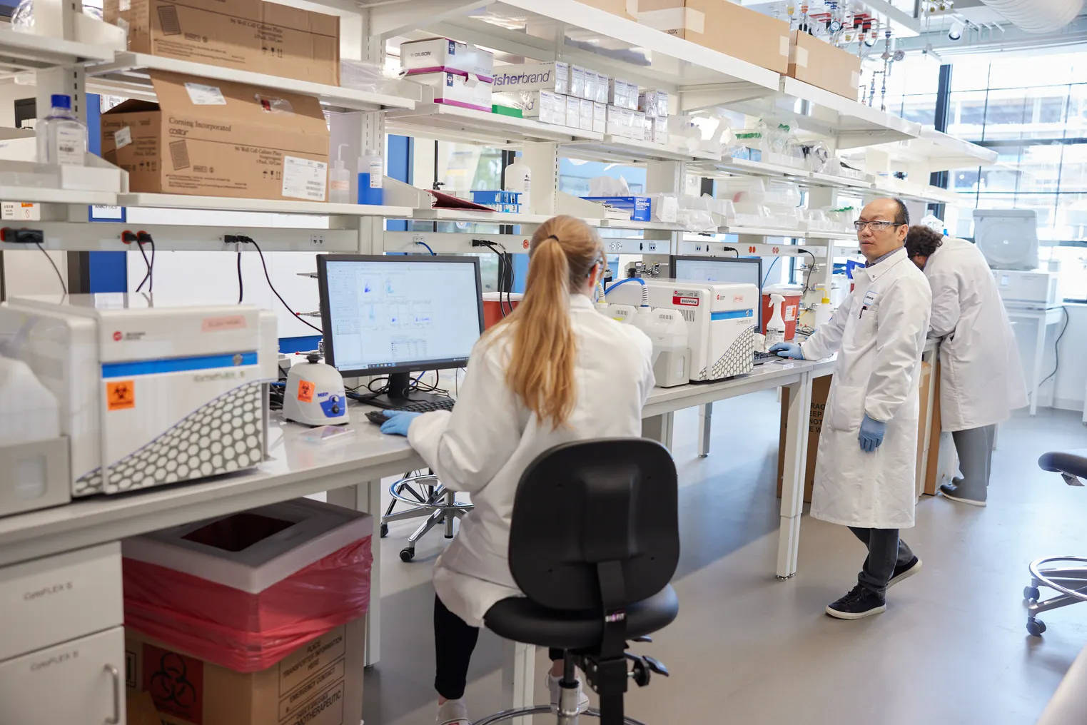 Two lab scientists work inside a research facility. A man turns to talk to his colleague, who is seated.