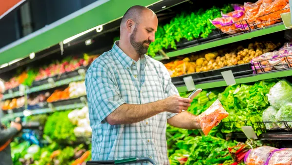 A shopper uses a phone while holding a bag of carrots inside a grocery store.