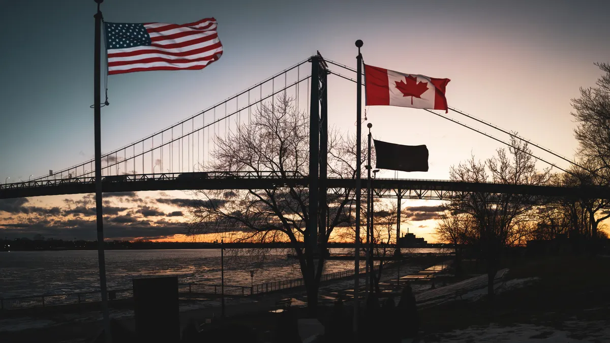 The Ambassador Bridge is shown with sunlight on the horizon and American and Canadian flags in the foreground.