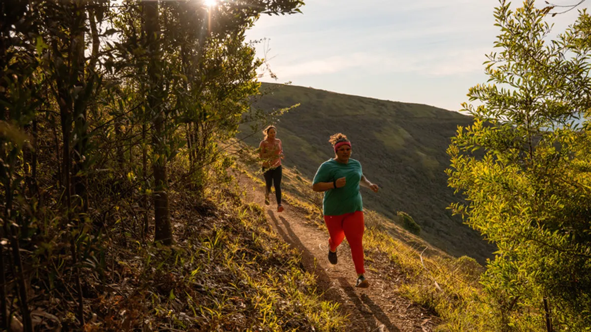 Two people run on a trail in a natural landscape.