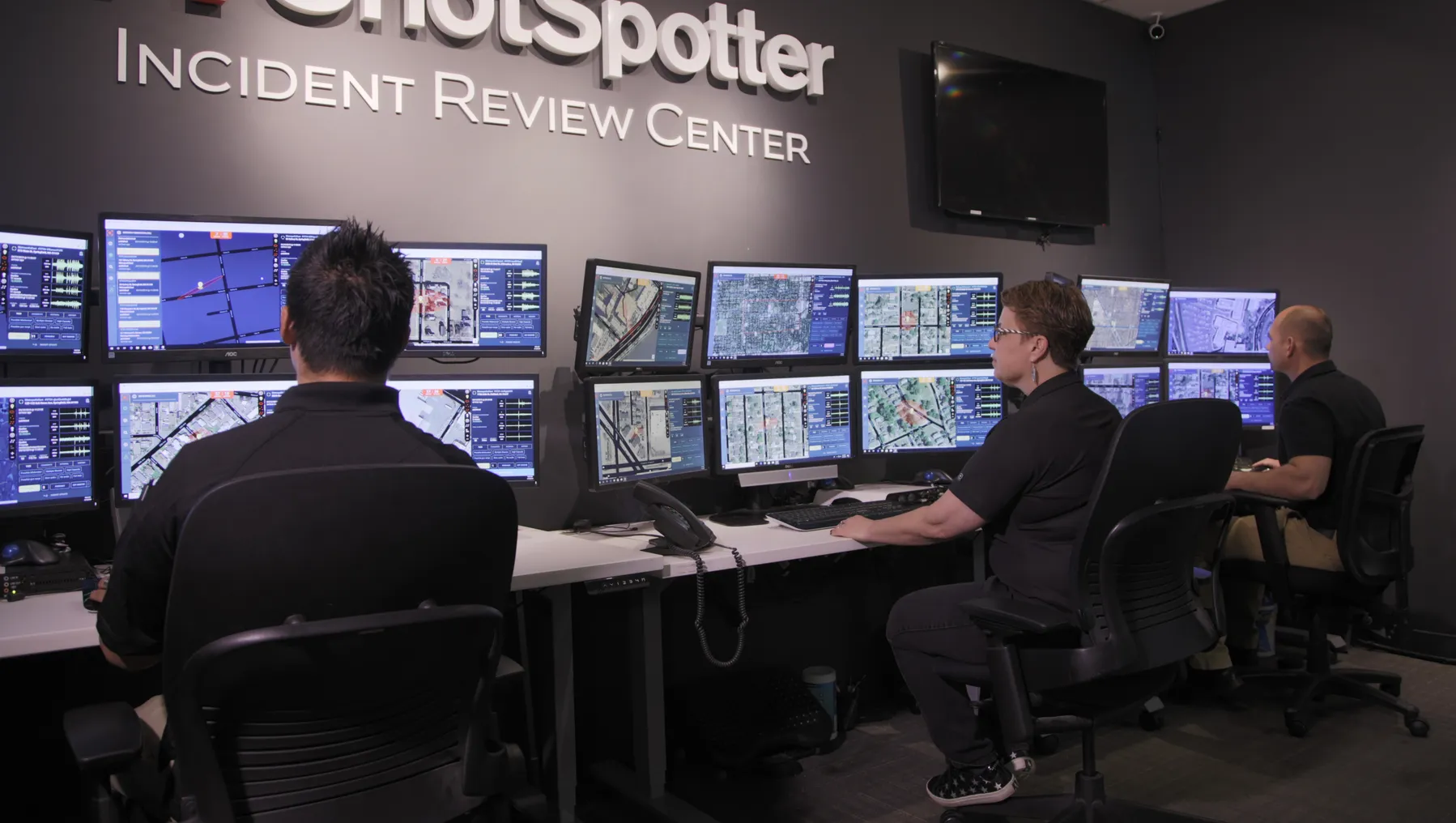 At ShotSpotter's Incident Review Center, three people sit in a row facing up to six computer monitors each, with keyboards and phones on the desks in front of them.