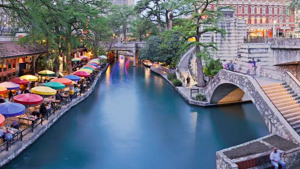 San Antonio's river walk area is shown, with the river in the center of the image, stone stairs on the right and a patio with colorful umbrellas on the left.