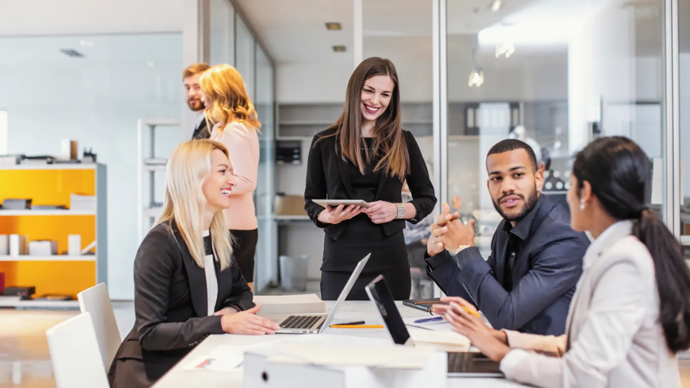 A diverse group of professionals in a modern office, engaged in a lively discussion around a table with laptops. The atmosphere is collaborative and positive.