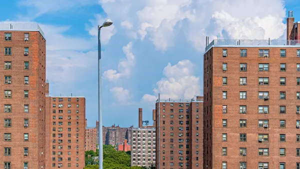A series of mid-rise apartment buildings characterized by their brick exteriors and regularly spaced windows reflect a cohesive architectural style, and trees run through the center opening of the buildings. 