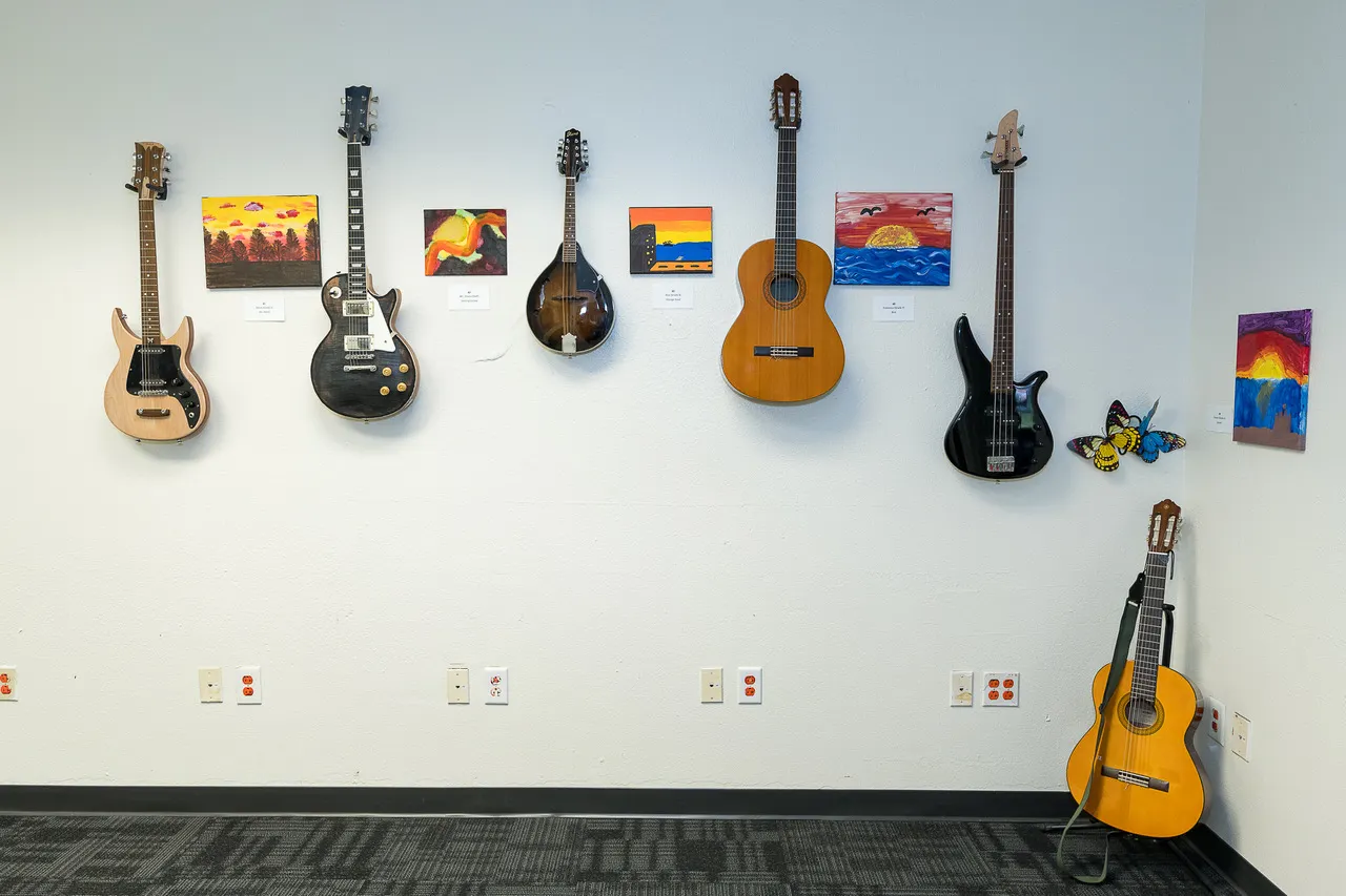 A variety of guitars and other string instruments adorn a white wall alongside student art in a high school's arts center.