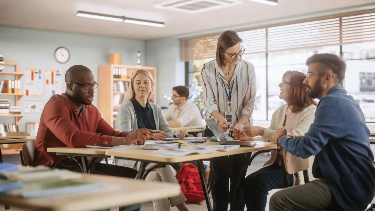 Several adults are seated around a table in a classroom. One person in the middle is standing and pointing to a sheet of paper on the table.