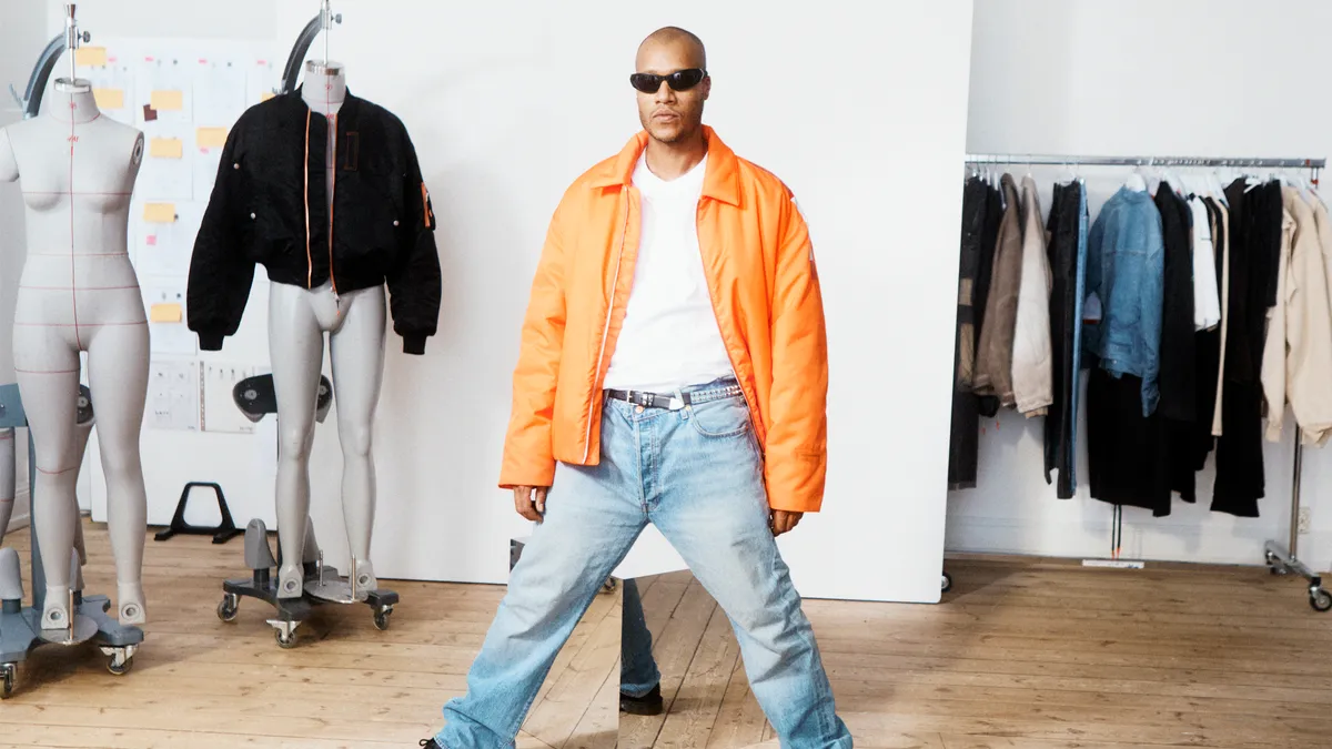 Heron Preston wears sunglasses and an orange jacket as he stands in an H&M workspace, surrounded by mannequins.