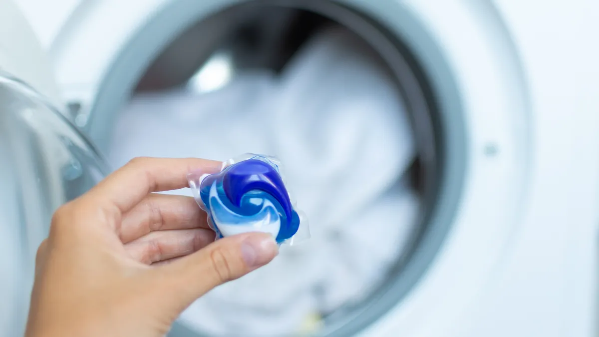 A person holds a blue and white laundry detergent pod in front of a washing machine.