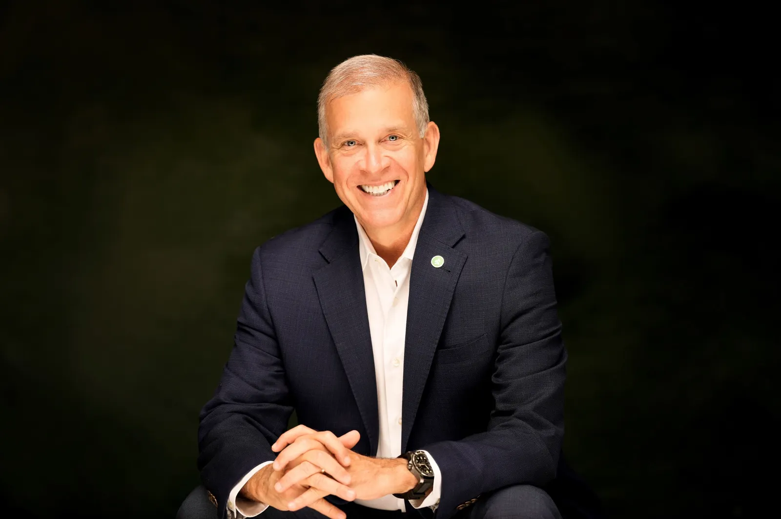 A person wearing a suit jacket sits with their hands clasped and smiles, in front of a black background.