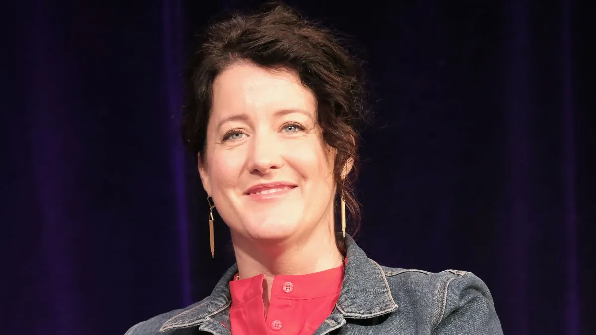 An image of a woman with brown hair wearing a jean jacket and red shirt in front of a dark blue curtain.