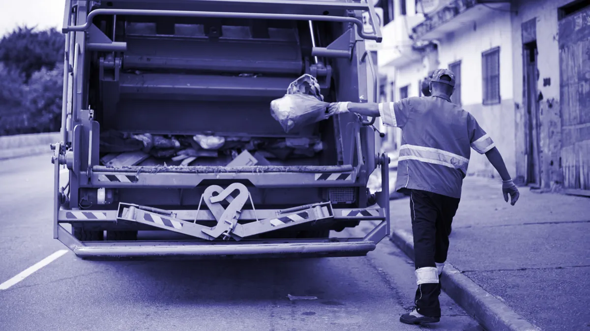 A person throws a bag of trash into a back of a waste collection truck on the street.