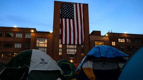 A view of the tents at George Washington University's encampment, with a large American flag draped over a campus building in the background.