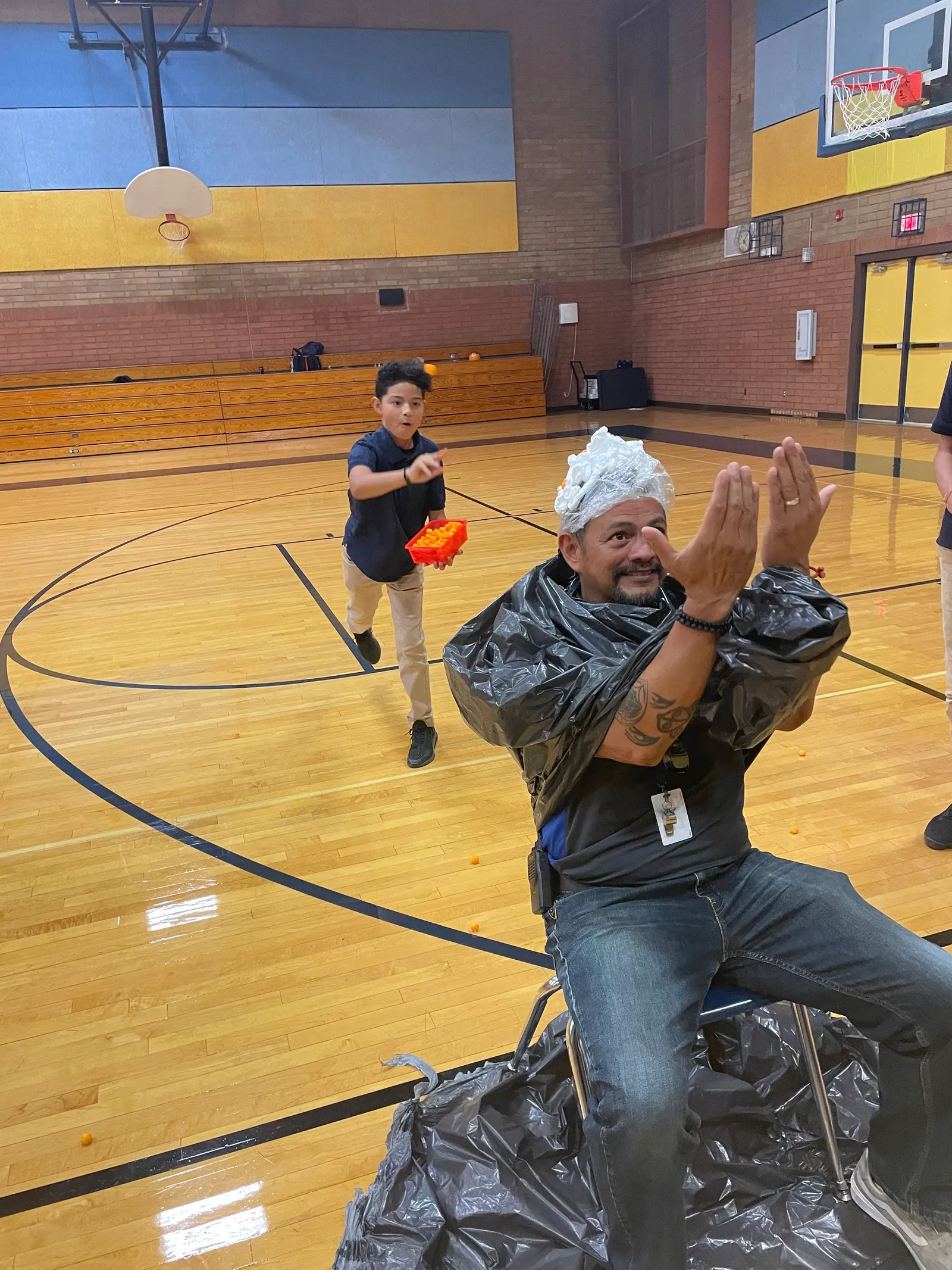 A man middle school principal with shaving cream on his head sits in a chair in a gymnasium while a student throws Cheeto puffs at his head.