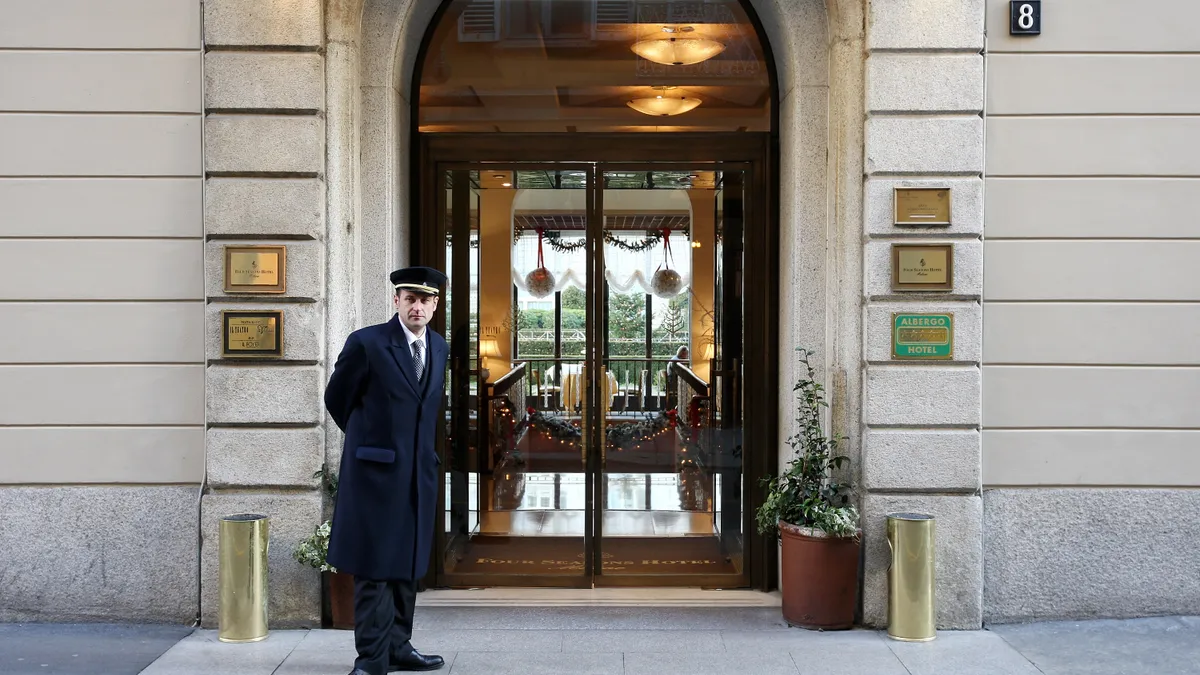 A doorman in a formal overcoat stands at the entrance of a hotel.