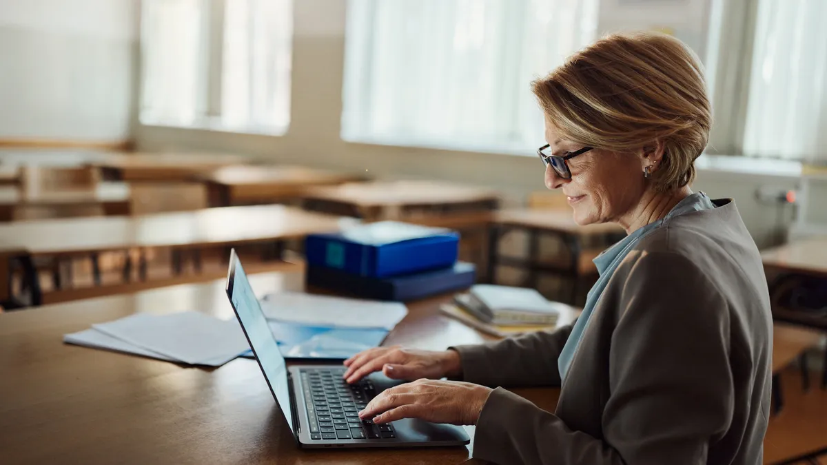 A teacher works alone at their desk while sitting in an empty classroom.