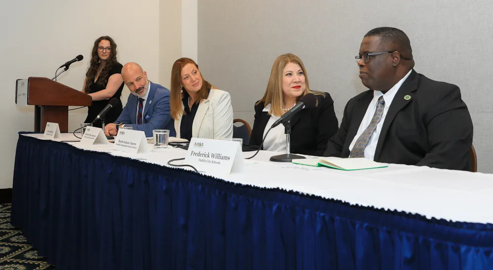 Four people are sitting at a table with microphones and name signs listening and talking. One additional person is standing at a podium.