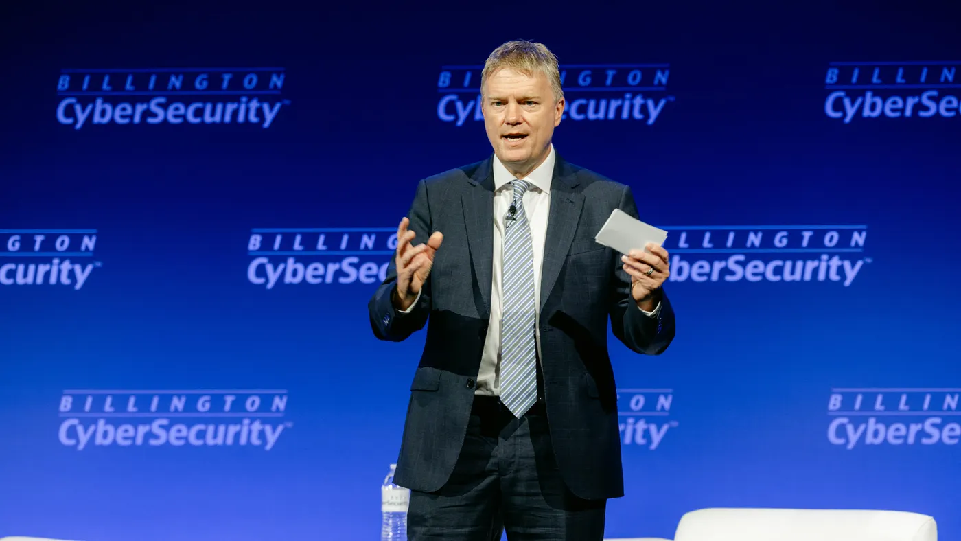 A person in business attire speaks while holding a card, with a blue background displaying the words "Billington Cybersecurity" in a tiled pattern.