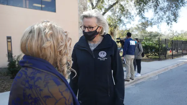 A person with white hair wearing black shirt and pants and a black mask talks to a person with blonde hair.