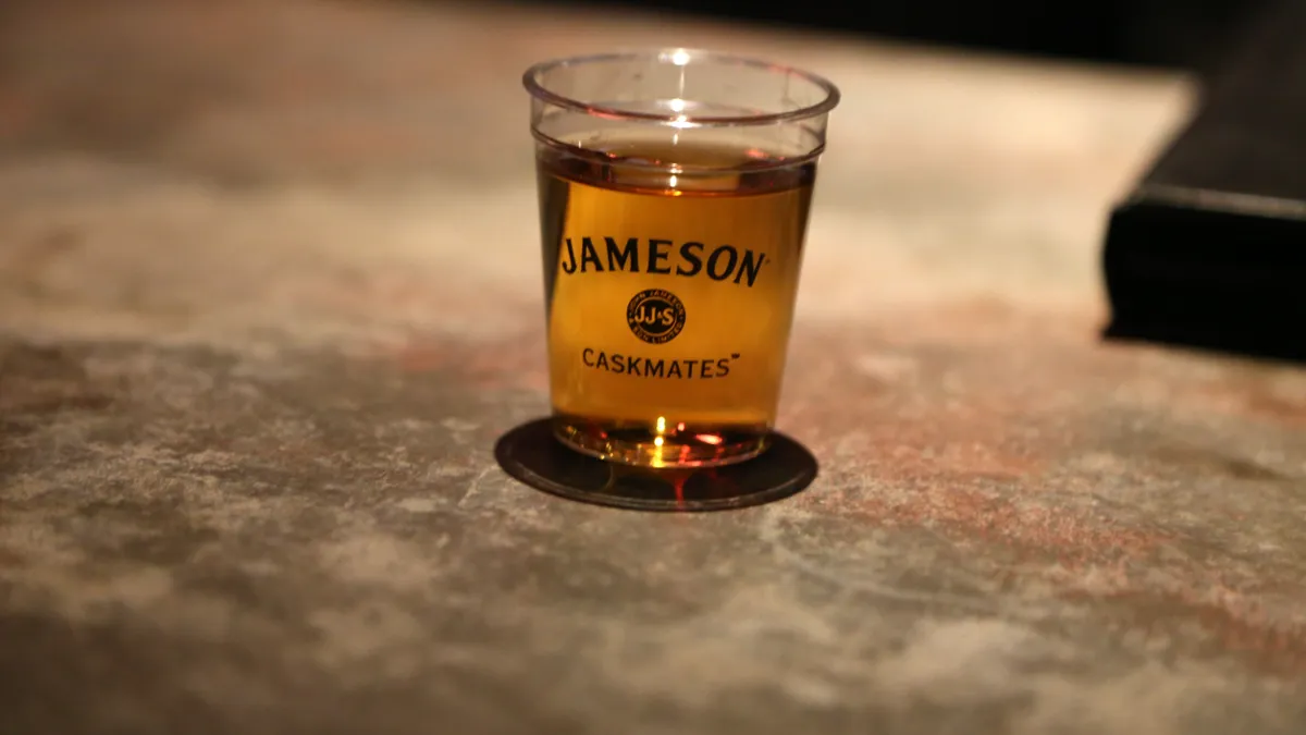 A dram of Jameson whiskey sits on a table in a branded cup.