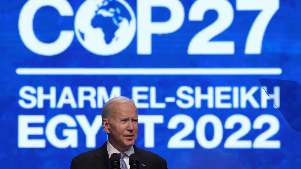 U.S. President Joe Biden stands on stage and speaks into a microphone at COP27 with the blue and white event logo and location on the screen behind him.