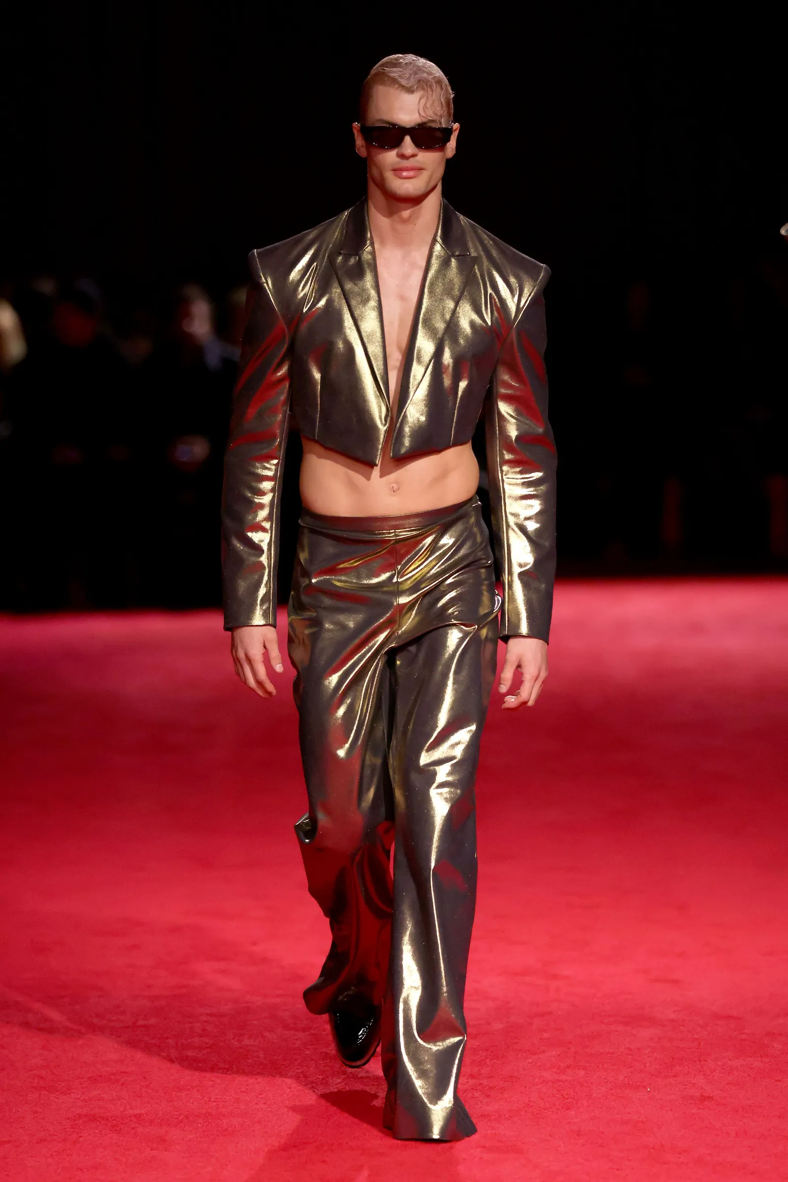 A model in a gold two-piece cropped suit walks down a runway.