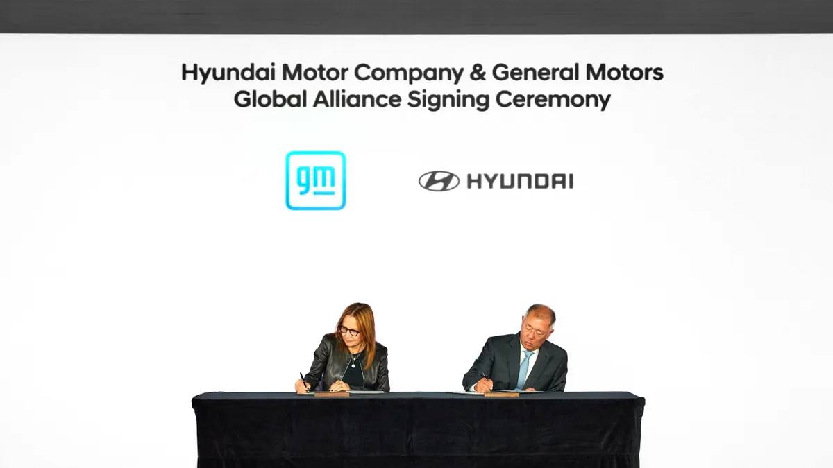 GM CEO Mary Barra and Hyundai Motor Group Executive Chair Euisun Chung  signing the collaboration agreement while seated at a table covered in a black table cloth.