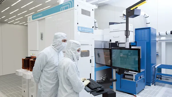 Two employees in PPE work on a computer beside Applied Materials equipment.